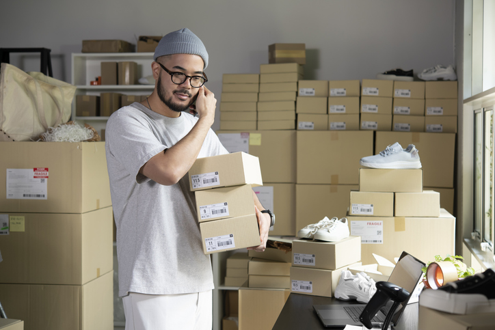 A man in a room full of boxes, making a phone call while holding a number of boxes.